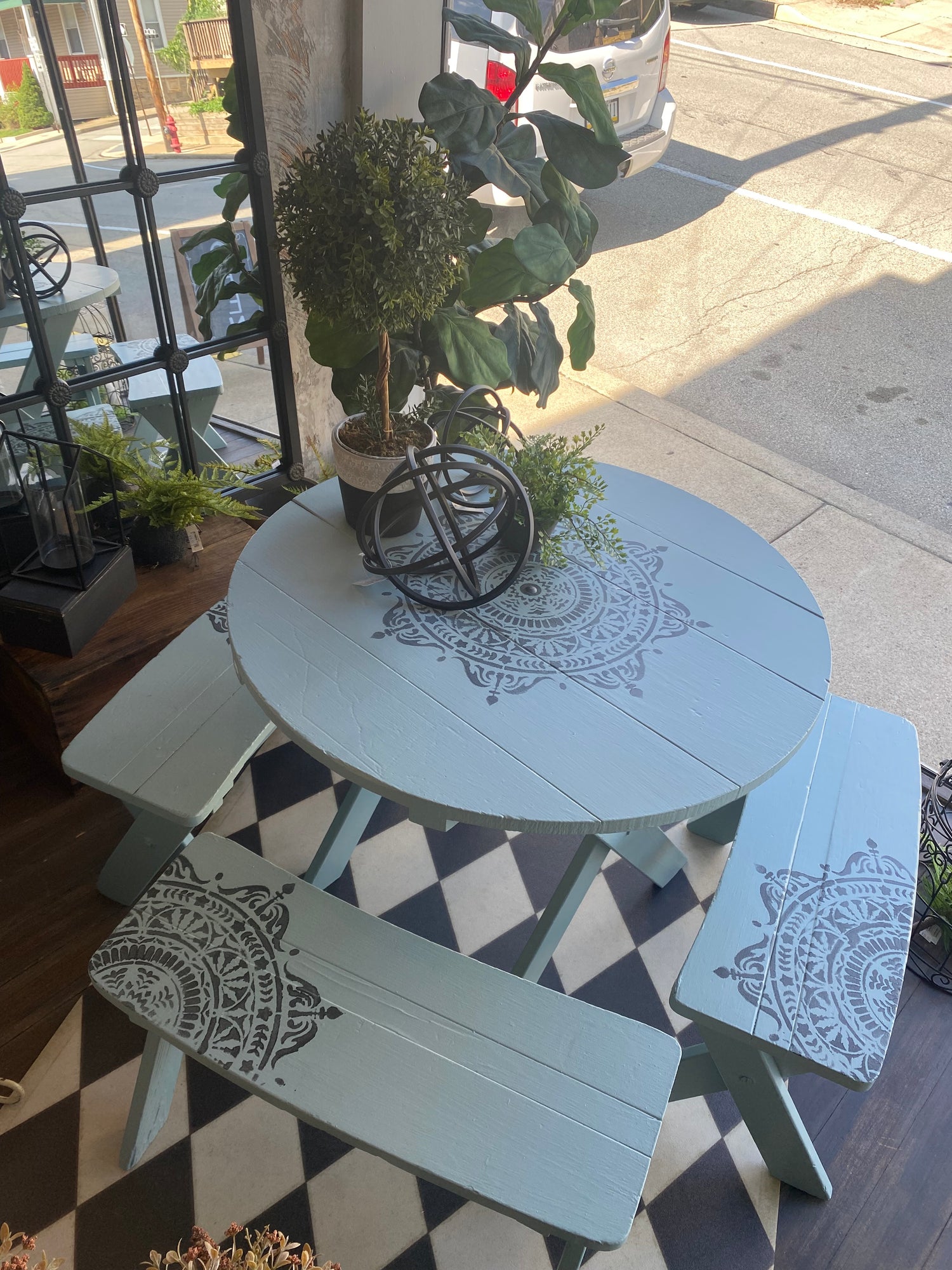 Image of a small round table sitting outside. The table is light gray with a darker gray design painted in the middle of the table and on each bench.